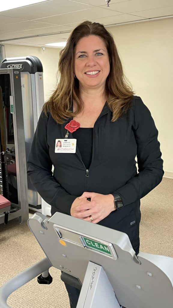 Jennifer is pictured smiling in black scrubs by workout machine.