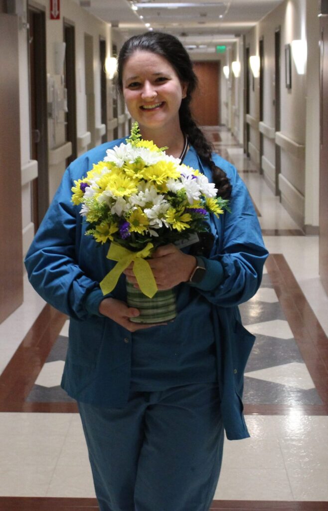 Kayla dressed in blue scrubs holding a beautiful white and yellow DAISY bouquet.