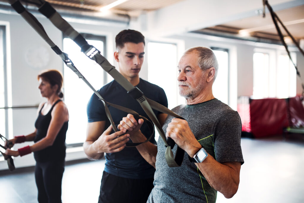 Senior man using TRX bands follows instructions from a young trainer. Personal trainers can help individuals create a customized plan for their fitness resolutions.