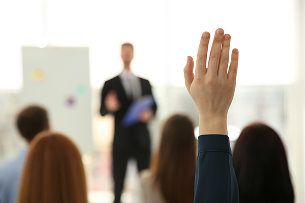 An individual is raising their hand while a man is speaking in a meeting.
