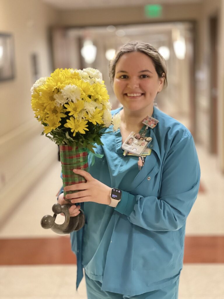 Nurse in blue scrubs, smiling, holding bouquet of daisies.