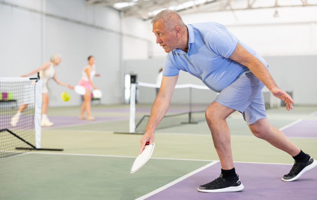 Older man plays pickleball on indoor court with other players in the background. The ability to move is a gift that shouldn't be taken for granted. 