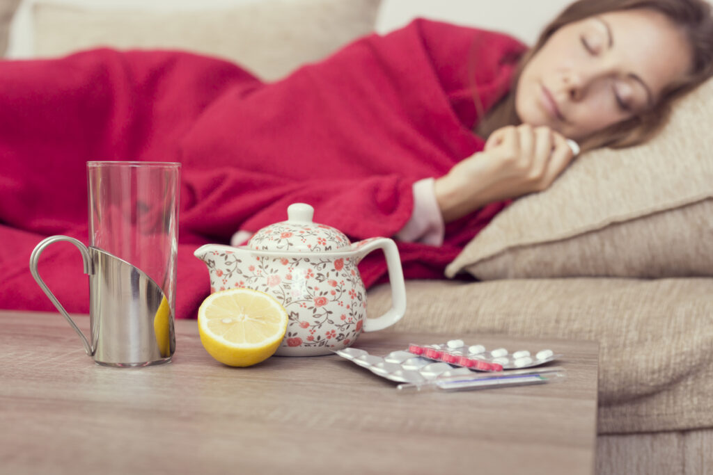 Wman resting on sofa; cold medicines, lemon, drinking glass and teapot are close by