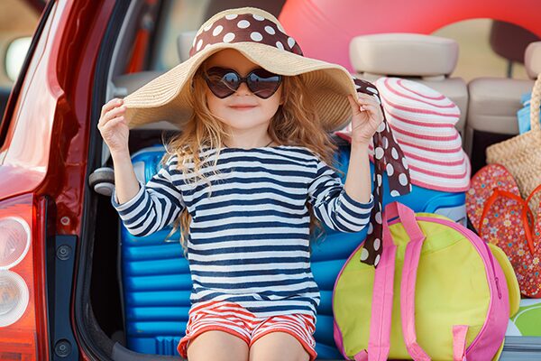 A little girl is sitting on the back of a car with her luggage packed and ready for vacation. 