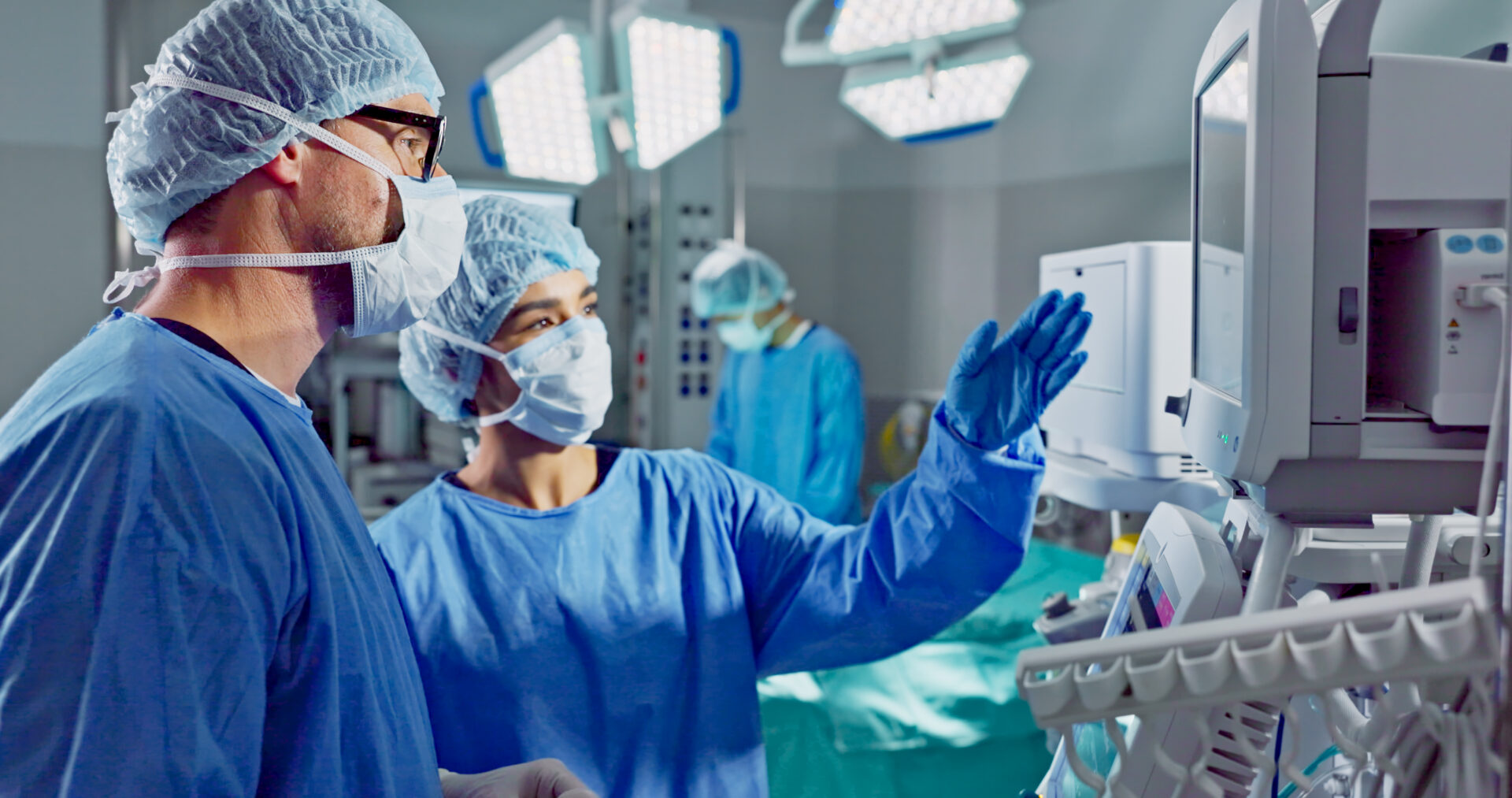 A male and female surgeon consult a monitor in a hospital operating room as they discuss a procedure.