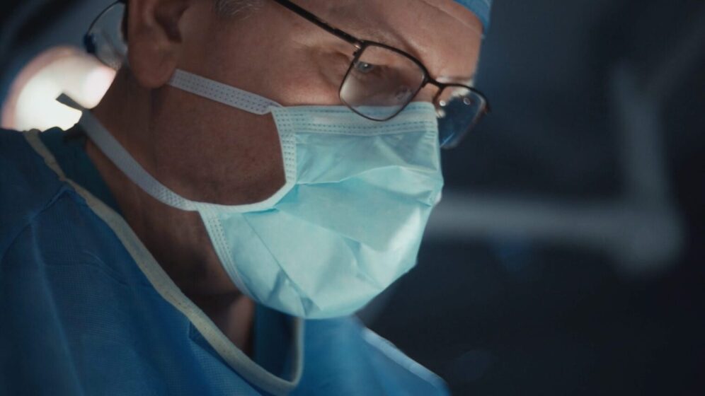 A male surgeon wearing a protective mask and glasses focuses on a surgical procedure.