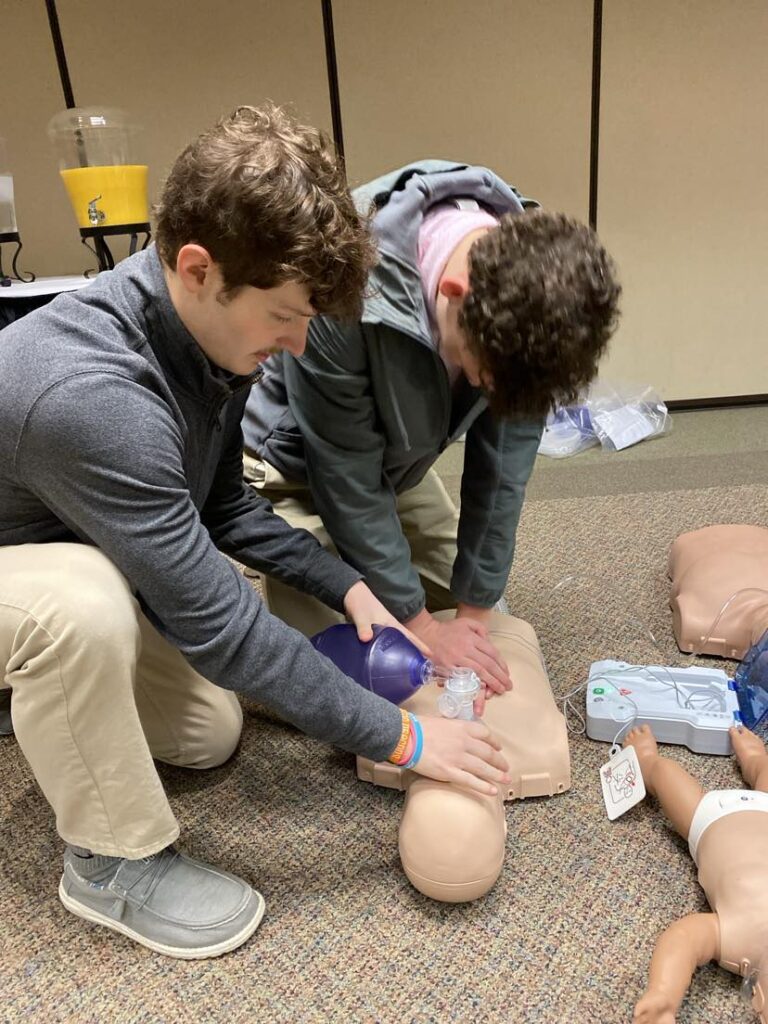 Two students work together to perform CPR on an adult mannequin.  One student performs compressions while one helps with breathing.