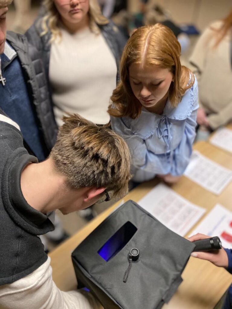 Students peer into a blacklight box to view residue left on hands after cleaning hands with either a hand sanitizing product or soap and water.